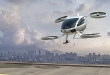 A generic white electric powered Vertical Take Off and Landing eVTOL aircraft with four rotors, coming in to land on roof top helipad with high city city buildings in the background. The sky is bright with clouds and it's late afternoon / early morning.