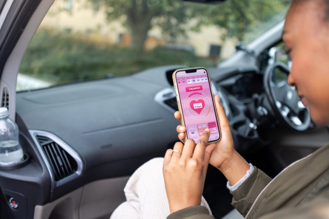 Over the shoulder view of a woman sitting in a van, using her mobile phone. She is smiling down at the screen in the North East of England. She is using a period tracking app, checking when her period is due.