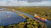 Container ship and car ferry navigate the Kiel Canal alongside vast solar panel fields and wind turbines, set against a backdrop of forest.