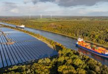 Container ship and car ferry navigate the Kiel Canal alongside vast solar panel fields and wind turbines, set against a backdrop of forest.