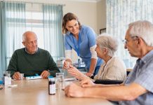 Young nurse in uniform giving medicine to group of seniors at retirement community. Happy smiling nurse gives medicine to elderly patients during her shift in a nursing home. Happy senior woman taking her dose of medicines from helpful caregiver at hospice.