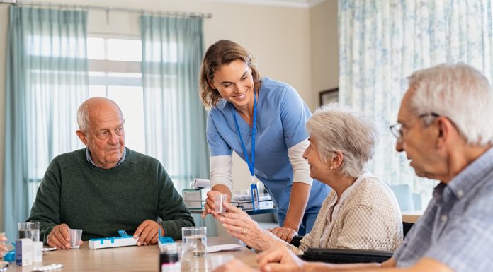 Young nurse in uniform giving medicine to group of seniors at retirement community. Happy smiling nurse gives medicine to elderly patients during her shift in a nursing home. Happy senior woman taking her dose of medicines from helpful caregiver at hospice.