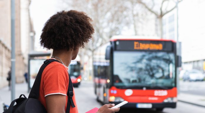 Side view of unrecognizable young African American man in casual clothes and backpack using smartphone while standing on city street and waiting for bus