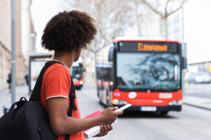Anonymous black male student waiting for bus on street Side view of unrecognizable young African American man in casual clothes and backpack using smartphone while standing on city street and waiting for bus