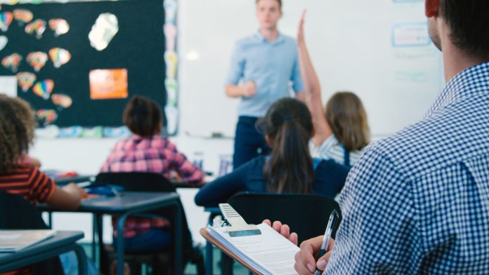 Male Trainee Teacher Being Observed Teaching In Classroom By Supervisor