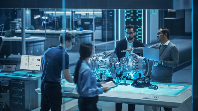 Young Team of Engineers, Project Manager and Machinery Operators Collaborate on a New Type of a Gas Turbine Engine, Standing with Tablet and Laptop Computers in Scientific Technology Lab. High Angle.