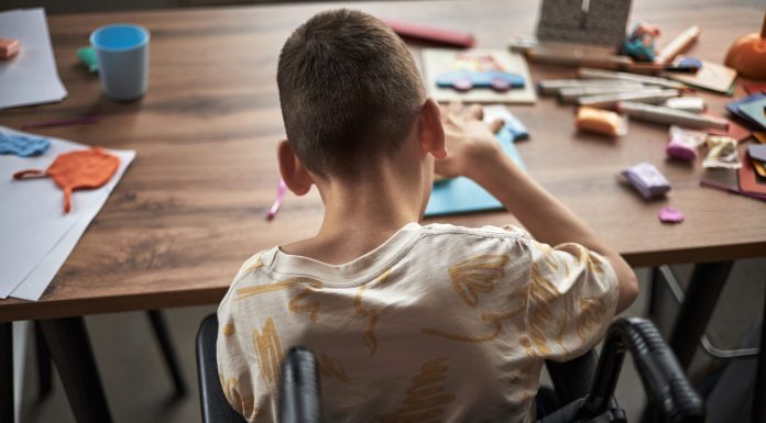 Child with disability engrossed in creating crafts while sitting at a cluttered table filled with art supplies and paper
