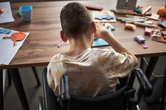 Child with disability engrossed in creating crafts while sitting at a cluttered table filled with art supplies and paper