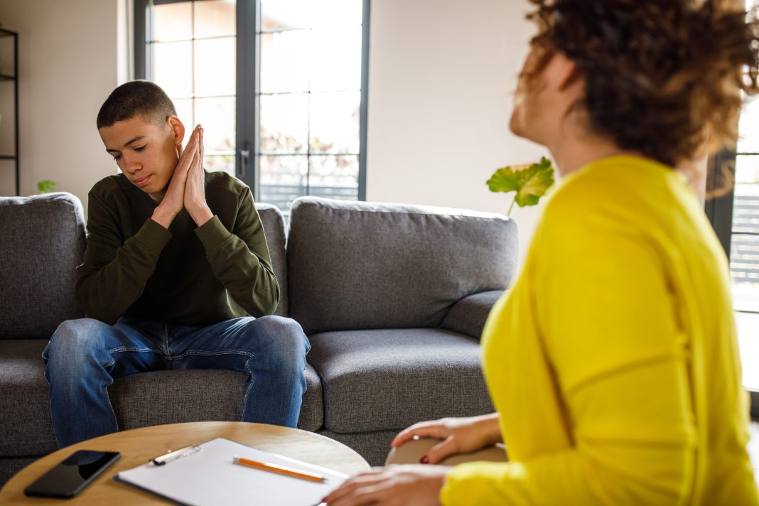 Selective focus shot of mid adult female therapist giving constructive advice to insecure teenage boy, that is struggling with negative emotions, sitting across from her on the sofa, during a therapy session.