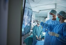 BMA slams Government’s ‘indefensible’ 2.5% pay offer for doctors Portrait of a Young Female and Male Surgeons Reviewing Patient's Scans on a Large Screen in a Dimly Lit Operating Room. Their Team of Assistants and Nurses Preparing for a Life-Saving Procedure