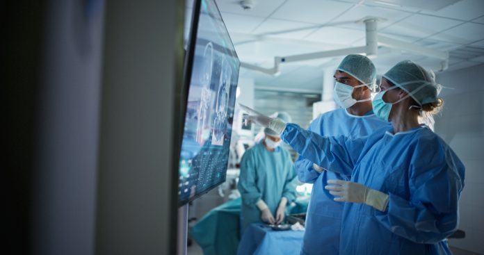 Portrait of a Young Female and Male Surgeons Reviewing Patient's Scans on a Large Screen in a Dimly Lit Operating Room. Their Team of Assistants and Nurses Preparing for a Life-Saving Procedure Portrait of a Young Female and Male Surgeons Reviewing Patient's Scans on a Large Screen in a Dimly Lit Operating Room. Their Team of Assistants and Nurses Preparing for a Life-Saving Procedure