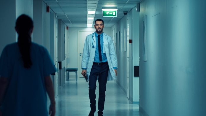 Determined Handsome Doctor Wearing White Coat with Stethoscope Walks Through Hospital Hallway. Modern Bright Clinic with Professional Staff.