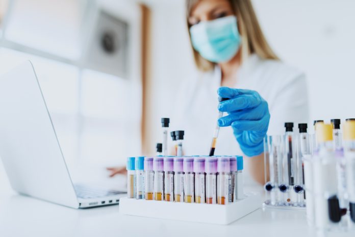 Close up of lab assistant in uniform, with mask and rubber gloves holding test tube with blood sample while sitting on chair and typing on laptop. Selective focus on test tubes. Close up of lab assistant in uniform, with mask and rubber gloves holding test tube with blood sample while sitting on chair and typing on laptop. Selective focus on test tubes.