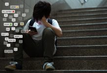 Asian teenage boy sitting at stair, covering his face with hands, face down, holding smartphone in low light.