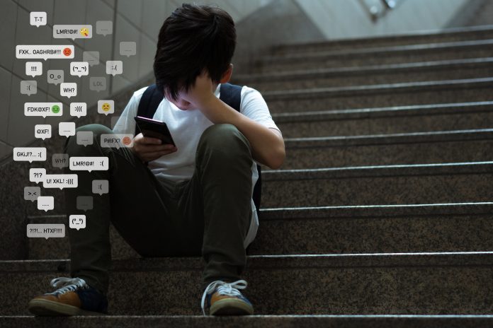 Asian teenage boy sitting at stair, covering his face with hands, face down, holding smartphone in low light.