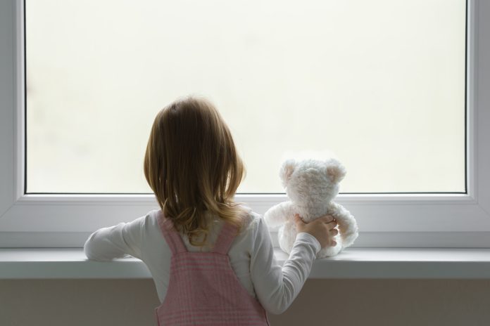 Little girl holding white teddy bear and standing alone at window and looking out from home. Back view. Waiting concept. Close up. Little girl holding white teddy bear and standing alone at window and looking out from home. Back view. Waiting concept. Close up.