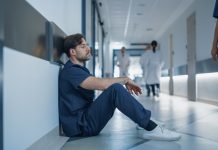 Young Stressed Male Doctor, Nurse or Surgeon in Scrubs Sitting Down Exhausted on a Hospital Hallway Floor, Trying to Rest, Fighting Stress and Fatigue in a Busy Medical Environment