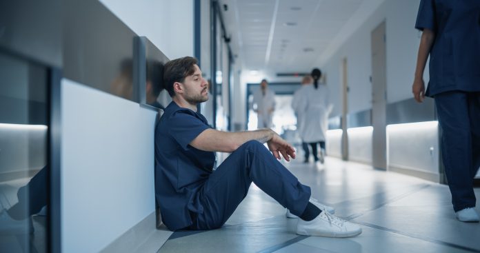Young Stressed Male Doctor, Nurse or Surgeon in Scrubs Sitting Down Exhausted on a Hospital Hallway Floor, Trying to Rest, Fighting Stress and Fatigue in a Busy Medical Environment Young Stressed Male Doctor, Nurse or Surgeon in Scrubs Sitting Down Exhausted on a Hospital Hallway Floor, Trying to Rest, Fighting Stress and Fatigue in a Busy Medical Environment