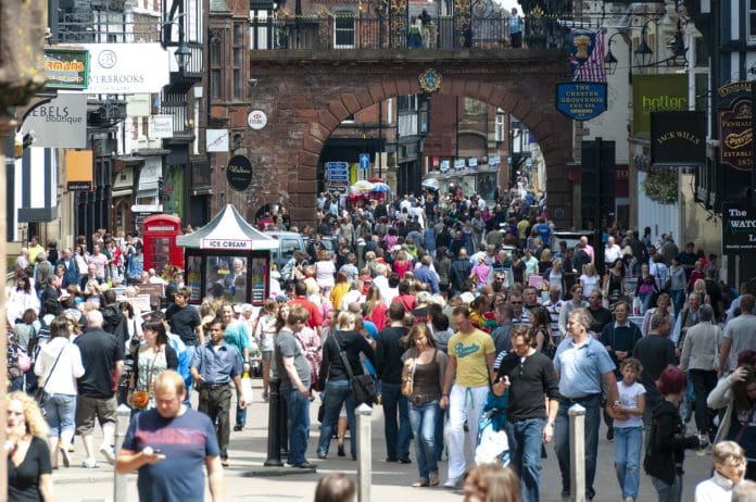 A vibrant scene of Chester High Street, bustling with shoppers and tourists. The street is lined with historic Tudor-style buildings, featuring distinctive black-and-white timber framing. People can be seen exploring the variety of shops, cafes, and local businesses that add to the lively atmosphere. The combination of charming architecture and a busy pedestrian area creates a dynamic and welcoming environment.